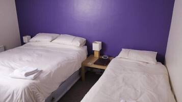 A hotel room with two beds, white bedding, a purple wall, a nightstand with a lamp, and a small table with a book and a remote control.