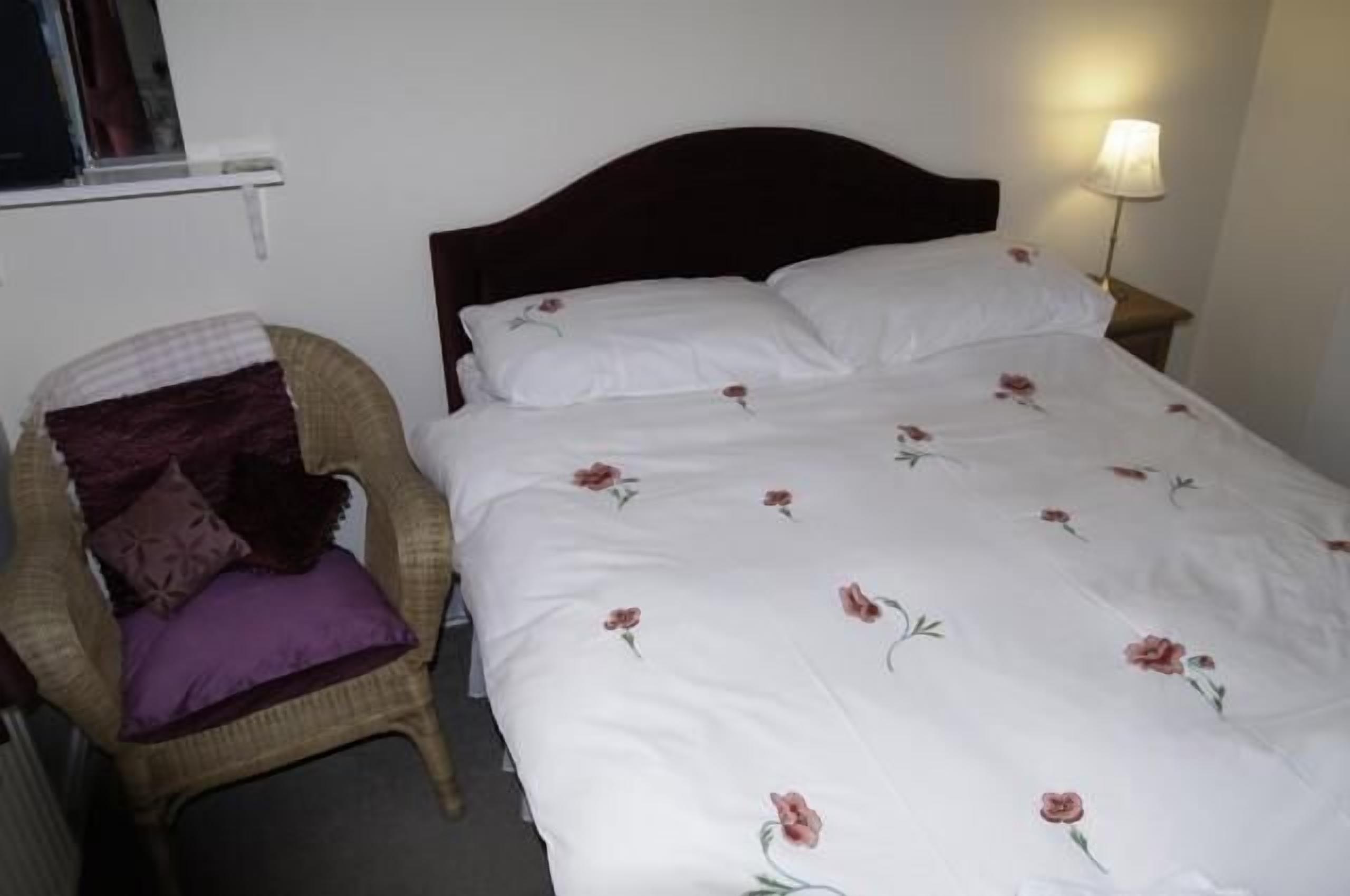 Een bed met een bloemenbedekking, een rieten stoel met een paars kussen en een bedlamp.