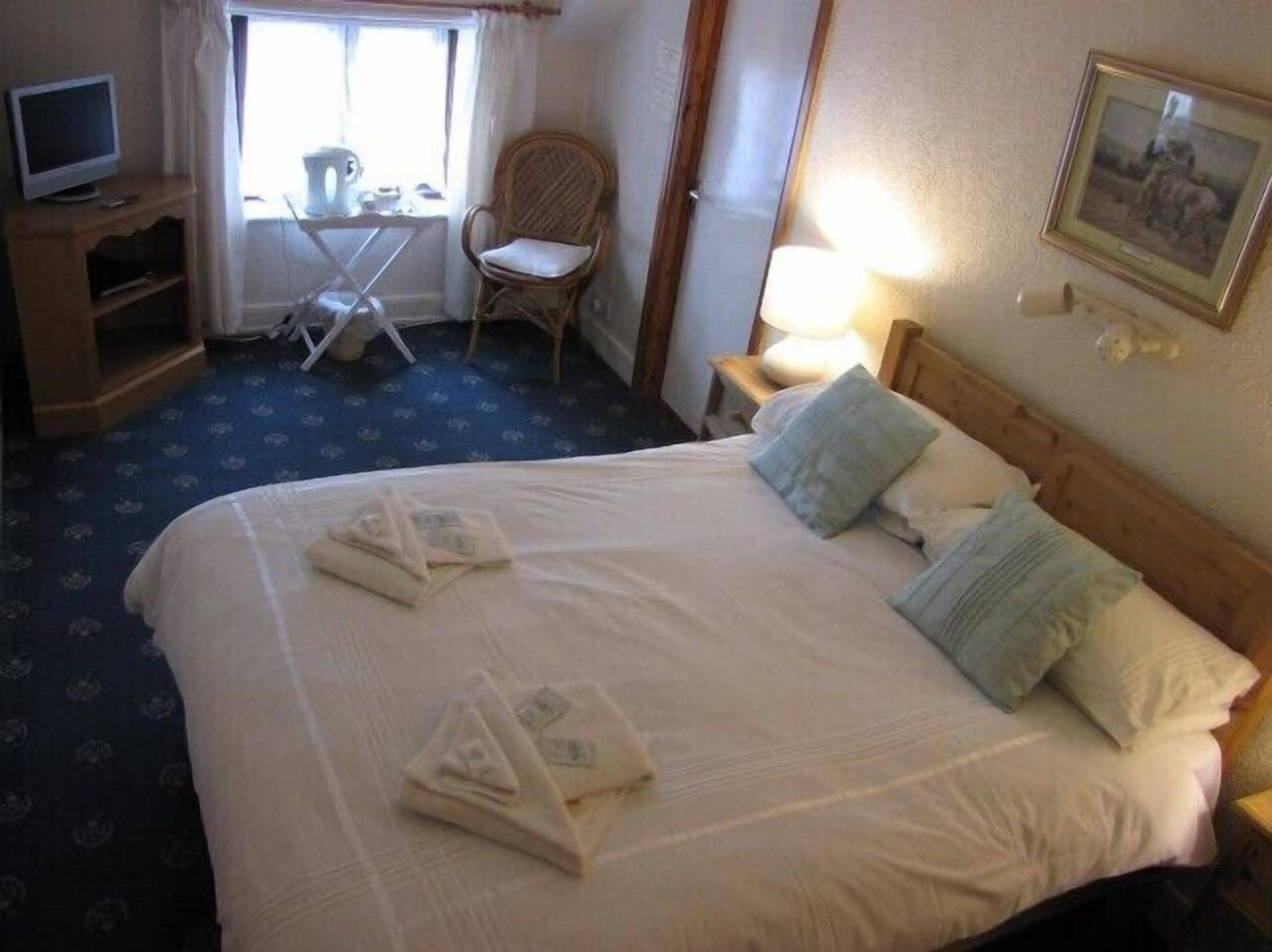 Habitación de hotel con una cama grande, un televisor en un soporte, una silla, una mesa y una ventana con cortinas.