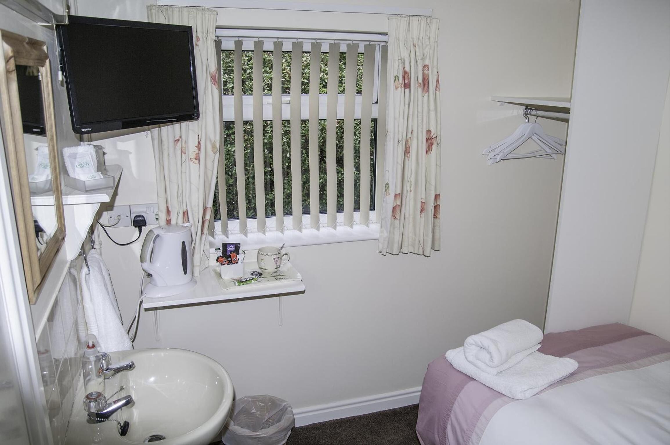 A small, clean room with a bed, a sink, a mirror, a television, and a window with curtains.