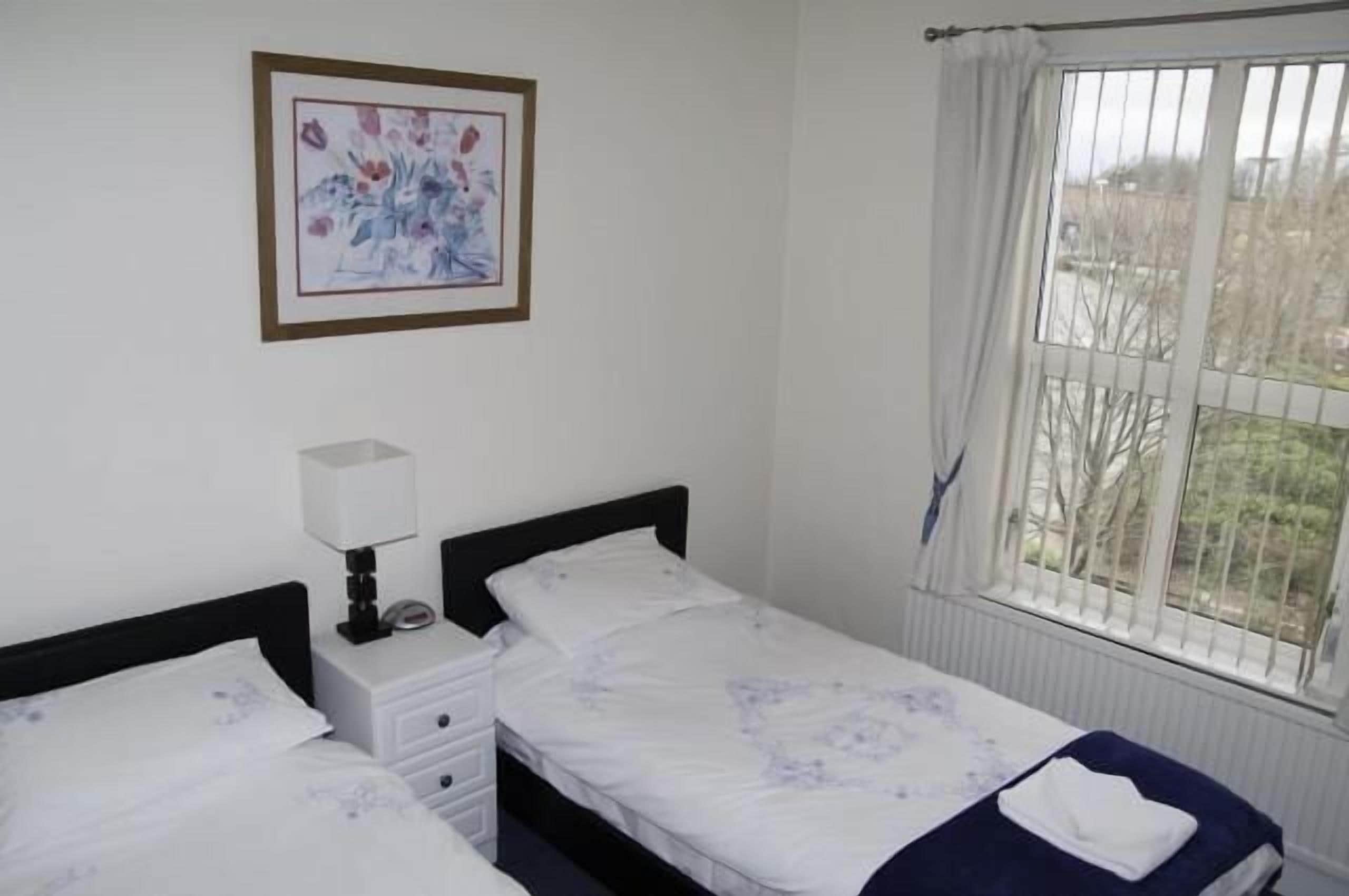A bedroom with two single beds, a bedside table with a lamp, a framed picture on the wall, and a window with curtains.