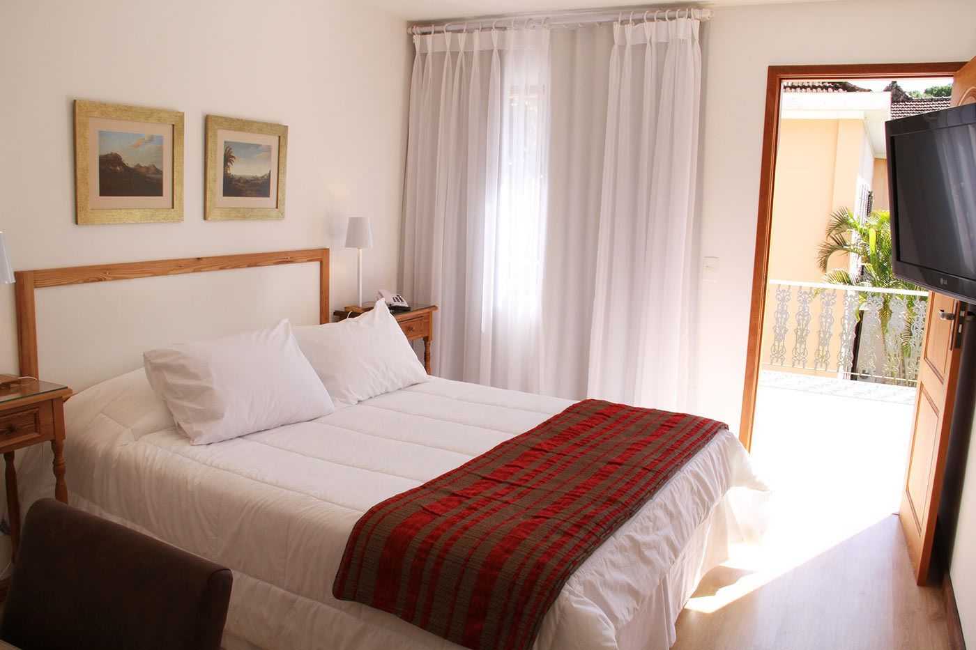 A hotel room with a bed, a nightstand, a lamp, two framed pictures on the wall, and a balcony with a TV.