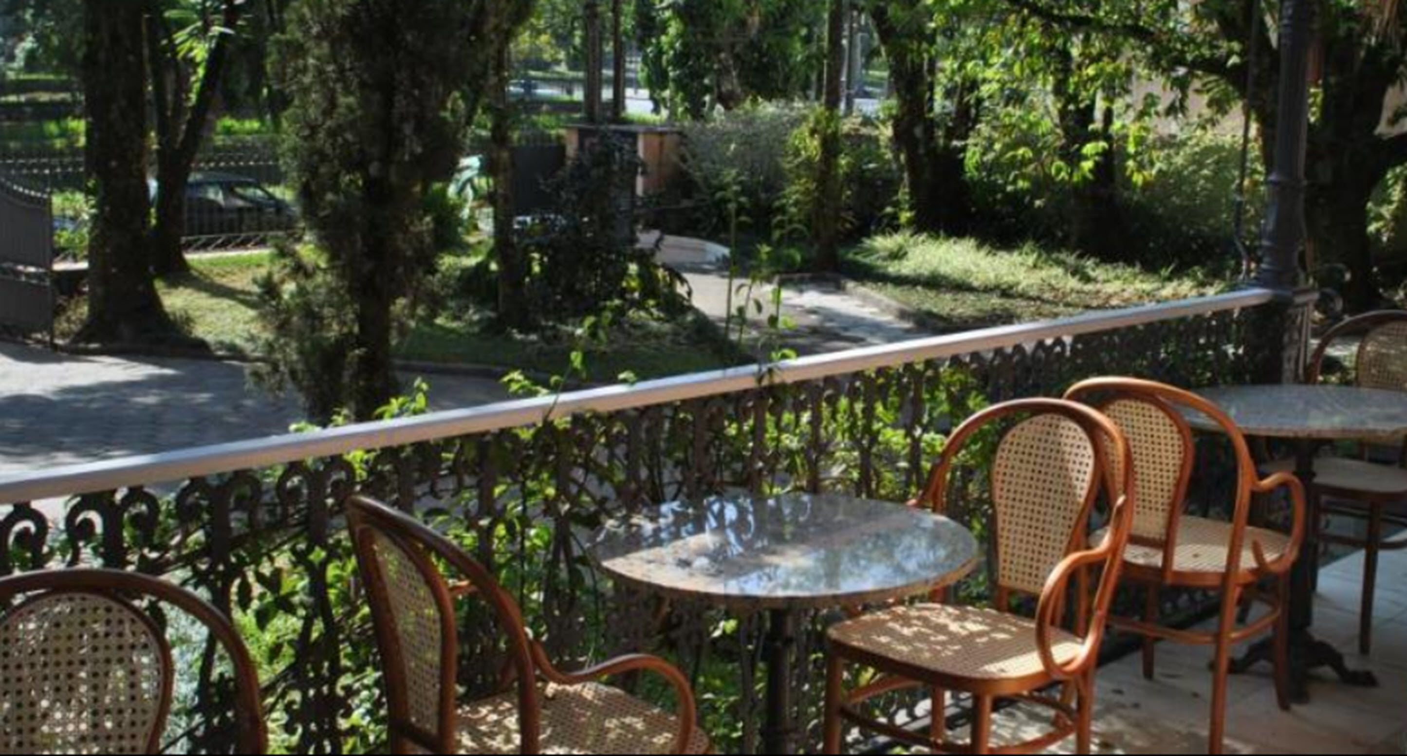 An outdoor patio with wicker chairs and a round table, surrounded by a metal railing and lush greenery.