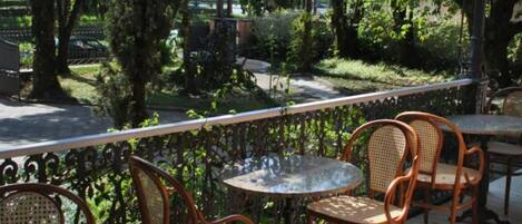 An outdoor patio with wicker chairs and a round table, surrounded by a metal railing and lush greenery.