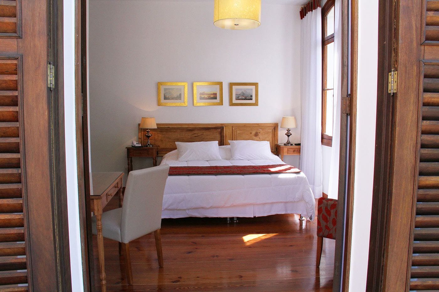 A bedroom with a wooden headboard, a bed with white linens, two bedside tables with lamps, and three framed pictures on the wall.