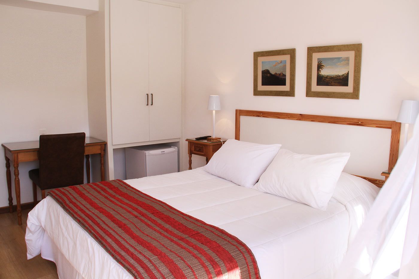 Una habitació d'hotel amb un llit, una taula de treball, una cadira, un armari i dos quadres penjats a la paret.
