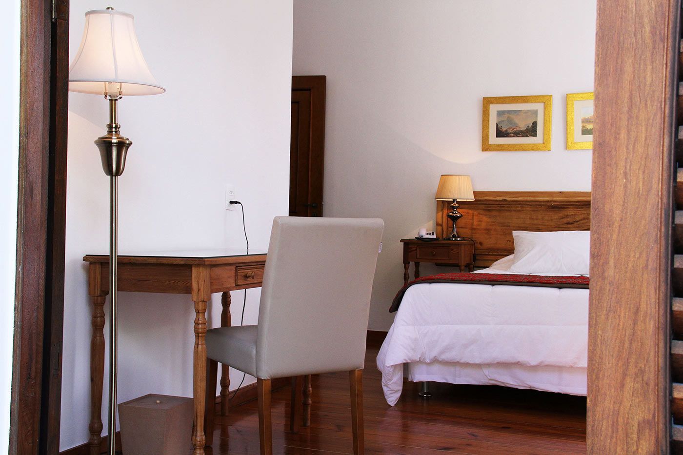 A hotel room with a wooden desk, a chair, a bed with a wooden headboard, a nightstand, a lamp, and two framed pictures on the wall.