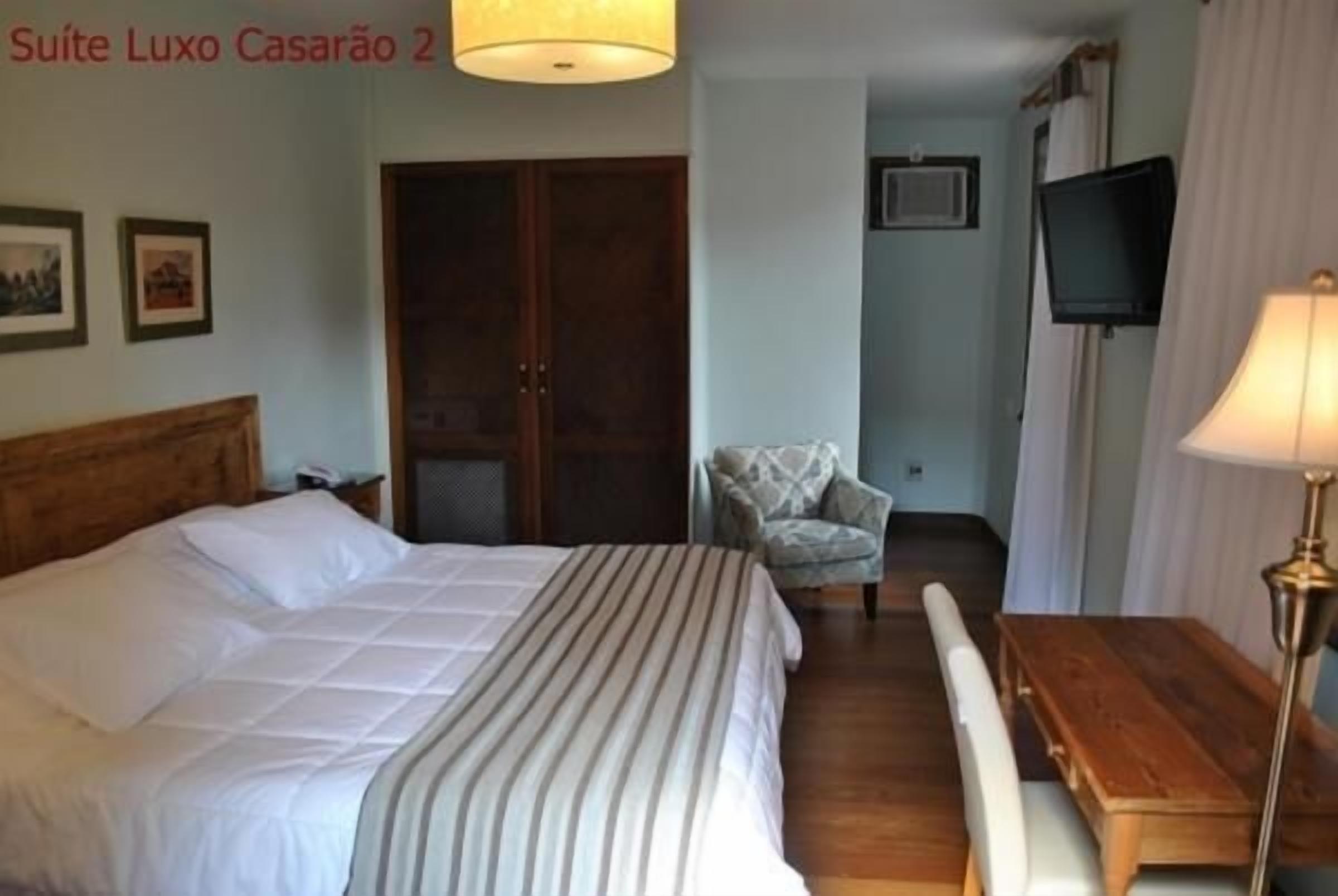 Habitación de hotel con cama, mesita de noche de madera, escritorio, silla, televisor y ventana con cortinas.