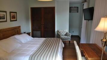 Habitación de hotel con cama, mesita de noche de madera, escritorio, silla, televisor y ventana con cortinas.