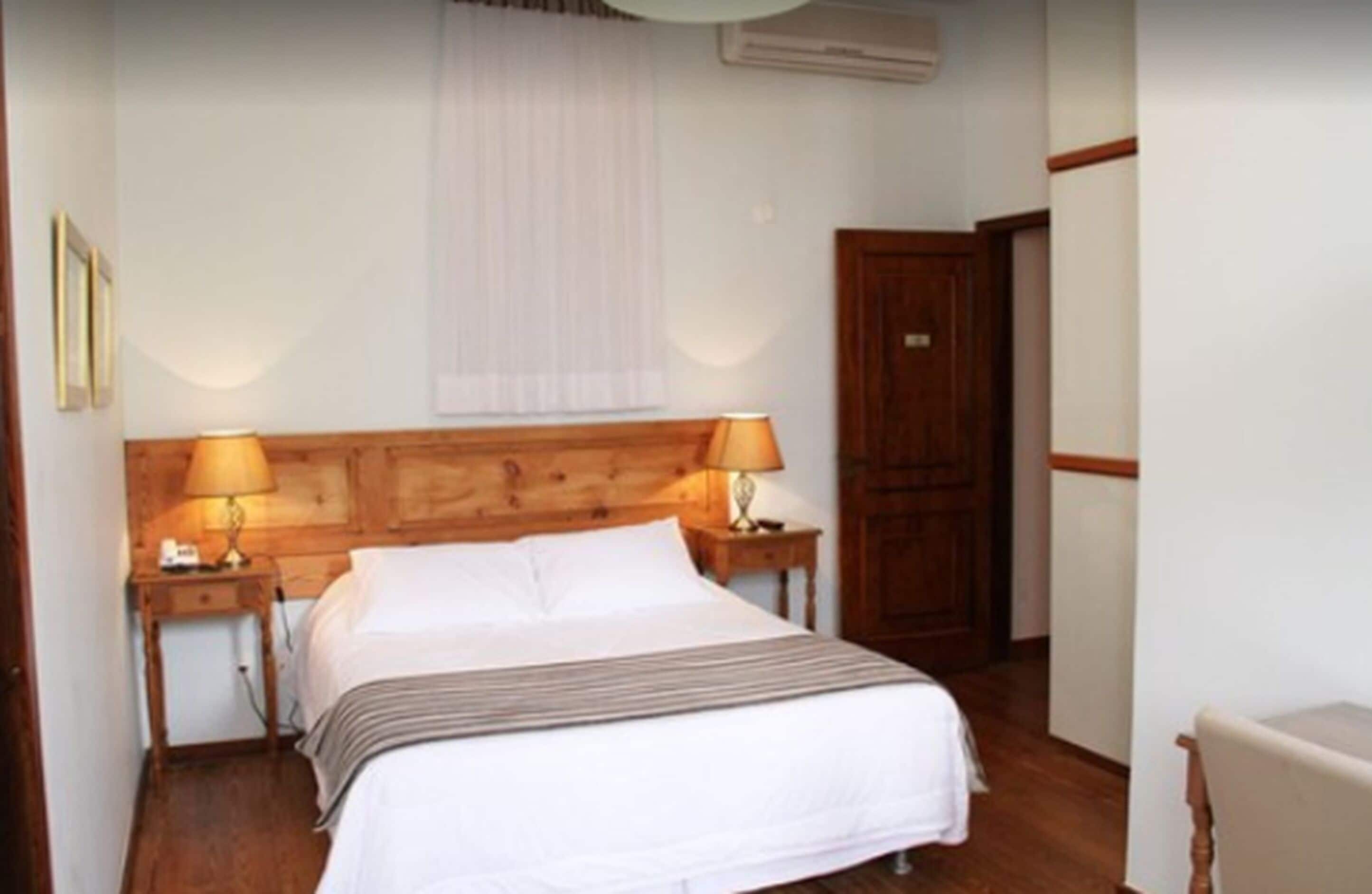 A neatly arranged bedroom with a wooden headboard, two bedside tables with lamps, a bed with white linens, a wooden wardrobe, and a door.