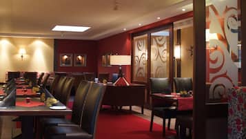 A hotel dining area with multiple tables and chairs, a red carpet, and wall-mounted lights.