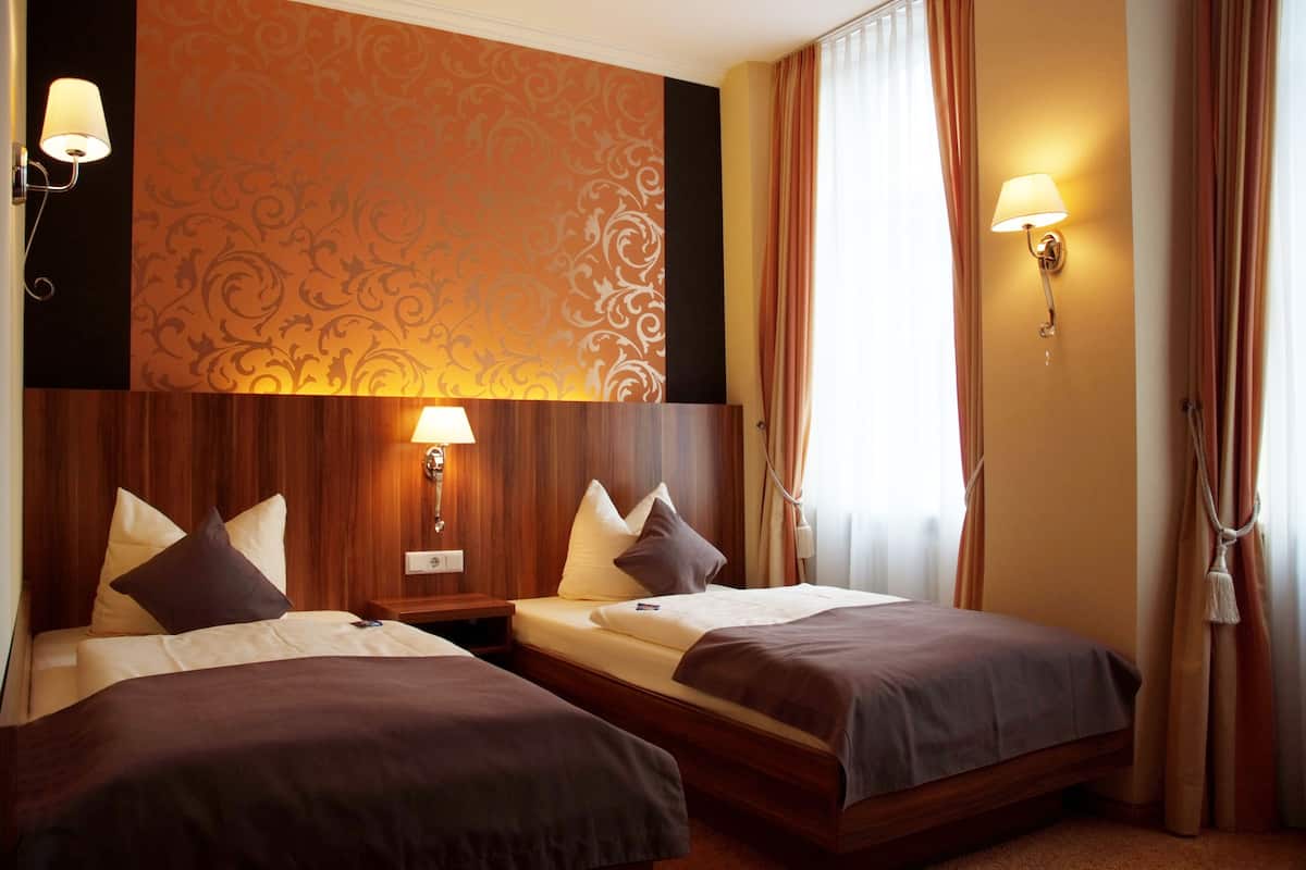 A hotel room with two single beds, wooden headboards, a patterned wall, and two wall-mounted lamps.
