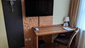Ett hotellrum med ett skrivbord i trä, en tv som är monterad på väggen, en stol, en papperskorg och ett fönster med gardiner.