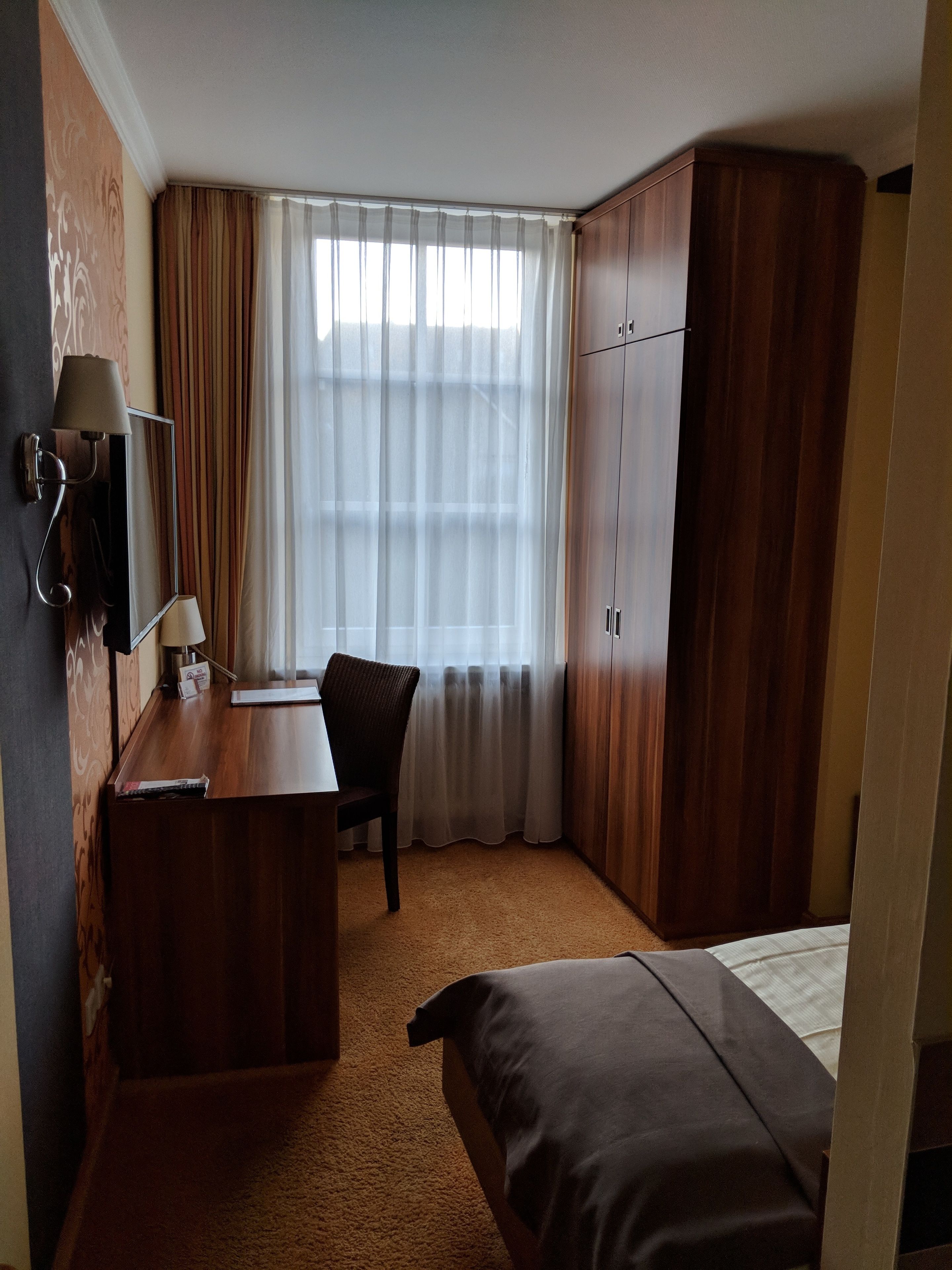 Una habitación de hotel con un escritorio de madera, una silla, una cama, un armario y una ventana con cortinas.