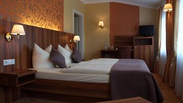 Una habitación de hotel con cabecera de madera, cama con muchos cojines, dos lámparas de noche, un televisor sobre un mueble de madera y una mesita.