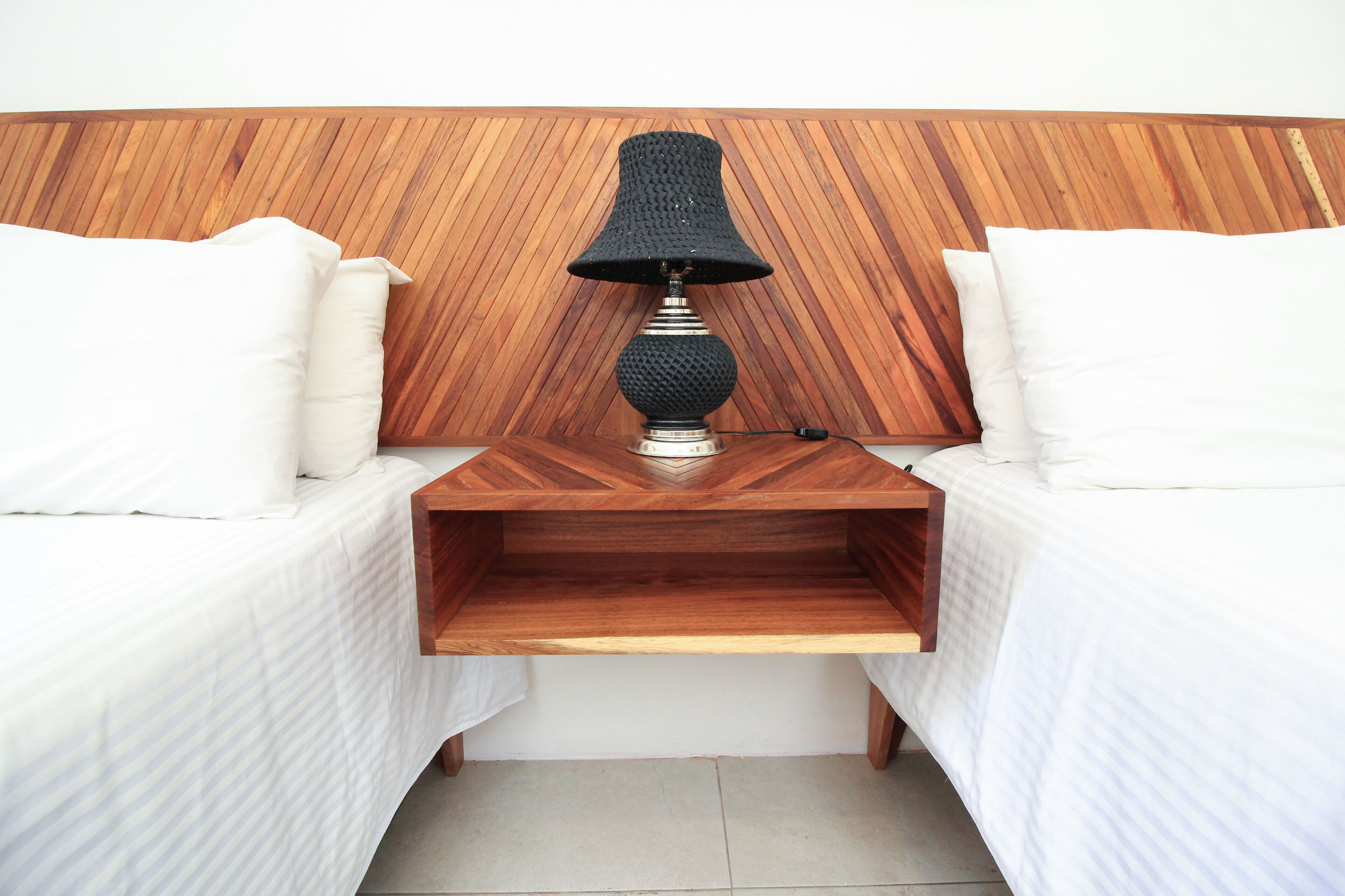 A bedroom with two beds, a wooden headboard, a bedside table with a lamp, and a small wooden shelf.