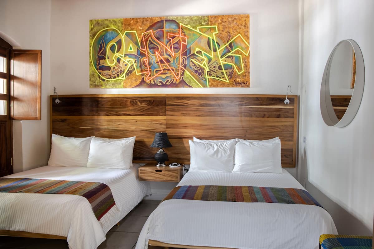 Habitación de hotel con dos camas, cabecera de madera, un cuadro abstracto de colores, una mesita de noche con lámpara y un espejo redondo en la pared.