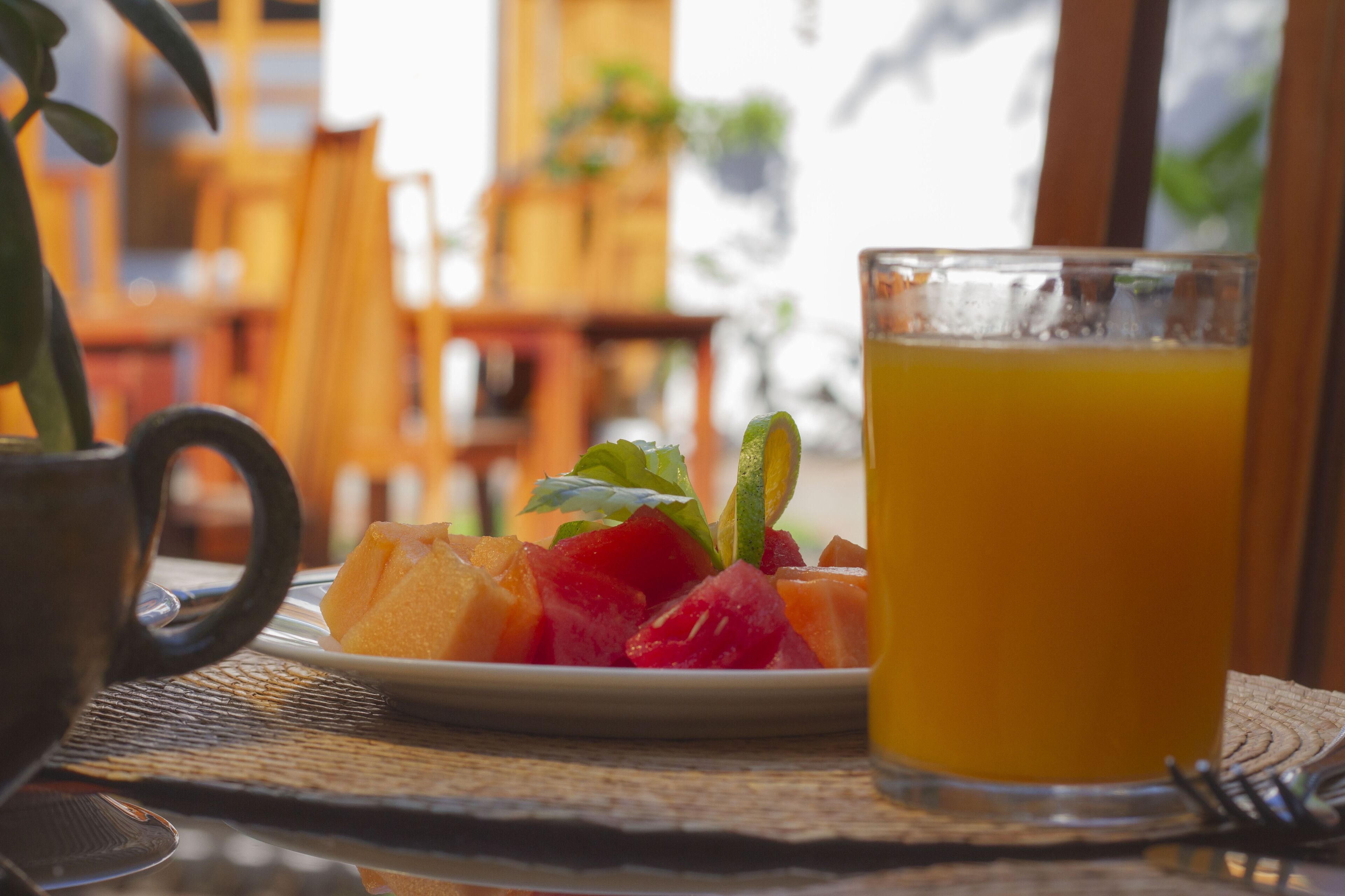 A table with a plate of fruit and a glass of orange juice, a coffee cup, and a fork.