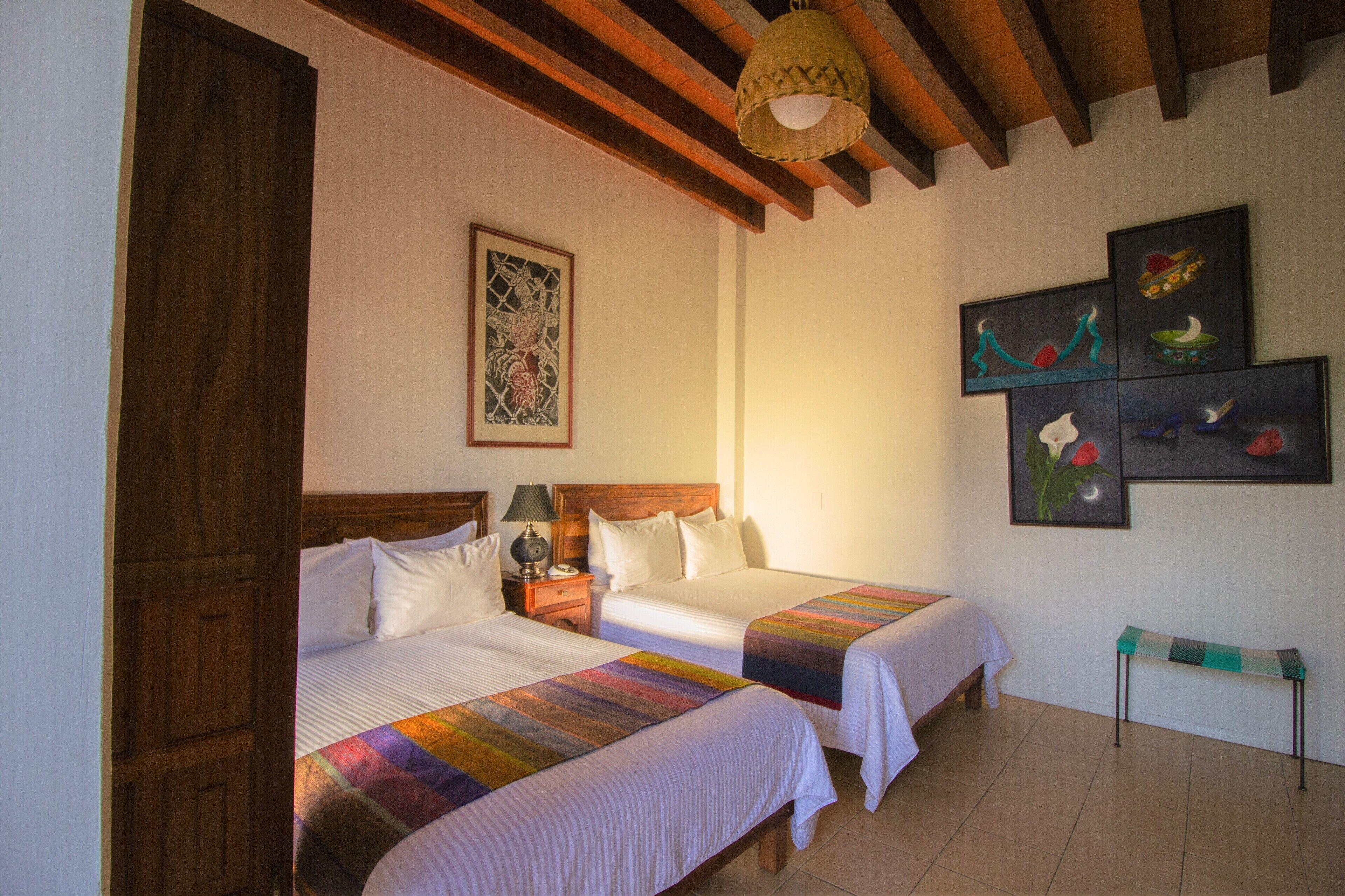 Habitación de hotel con dos camas, cabeceros de madera, una mesita de noche con lámpara, un banco y una obra de arte en la pared.