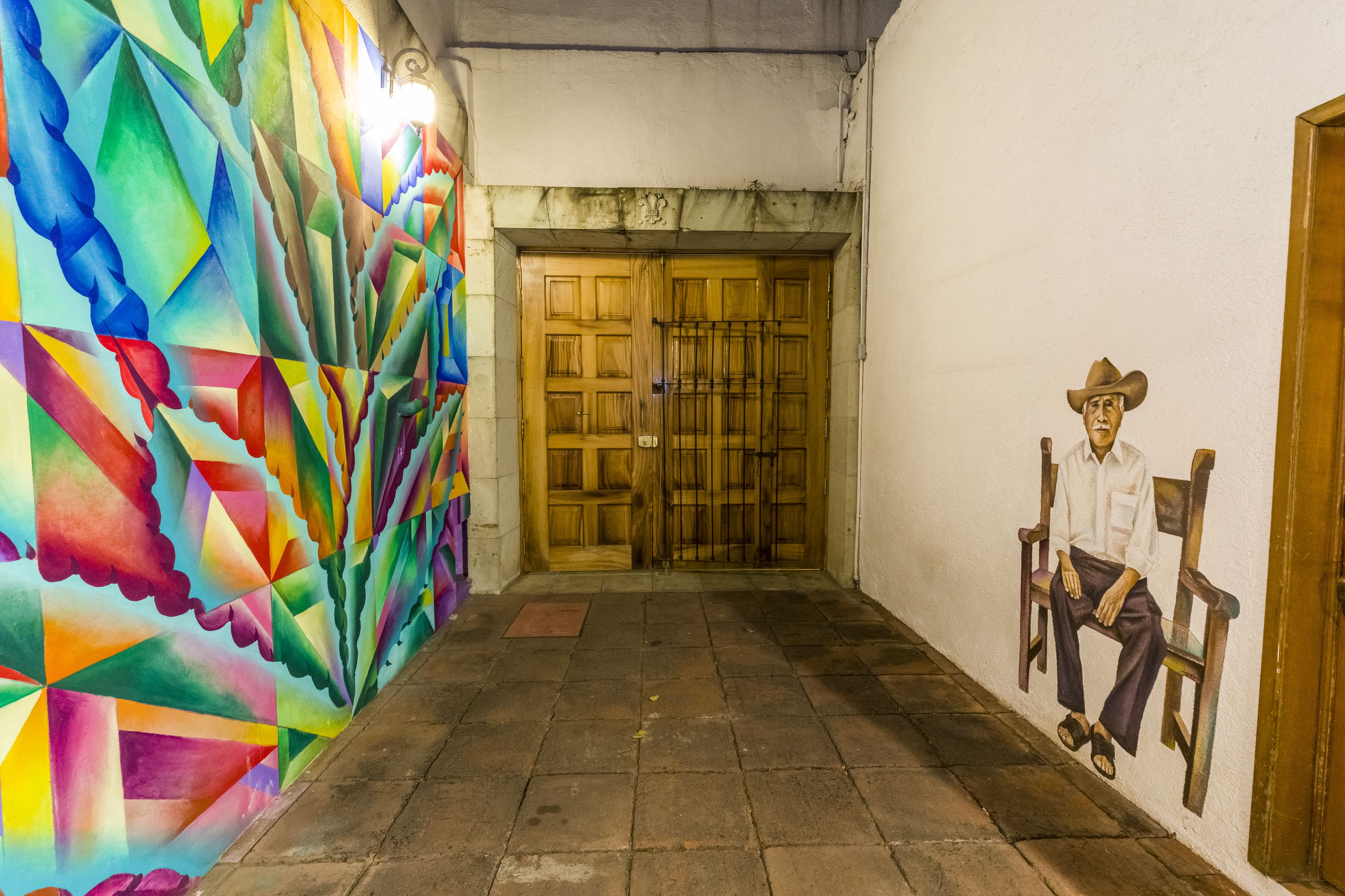 Un pasillo con un mural geométrico de colores a la izquierda y un mural de una figura sentada a la derecha.