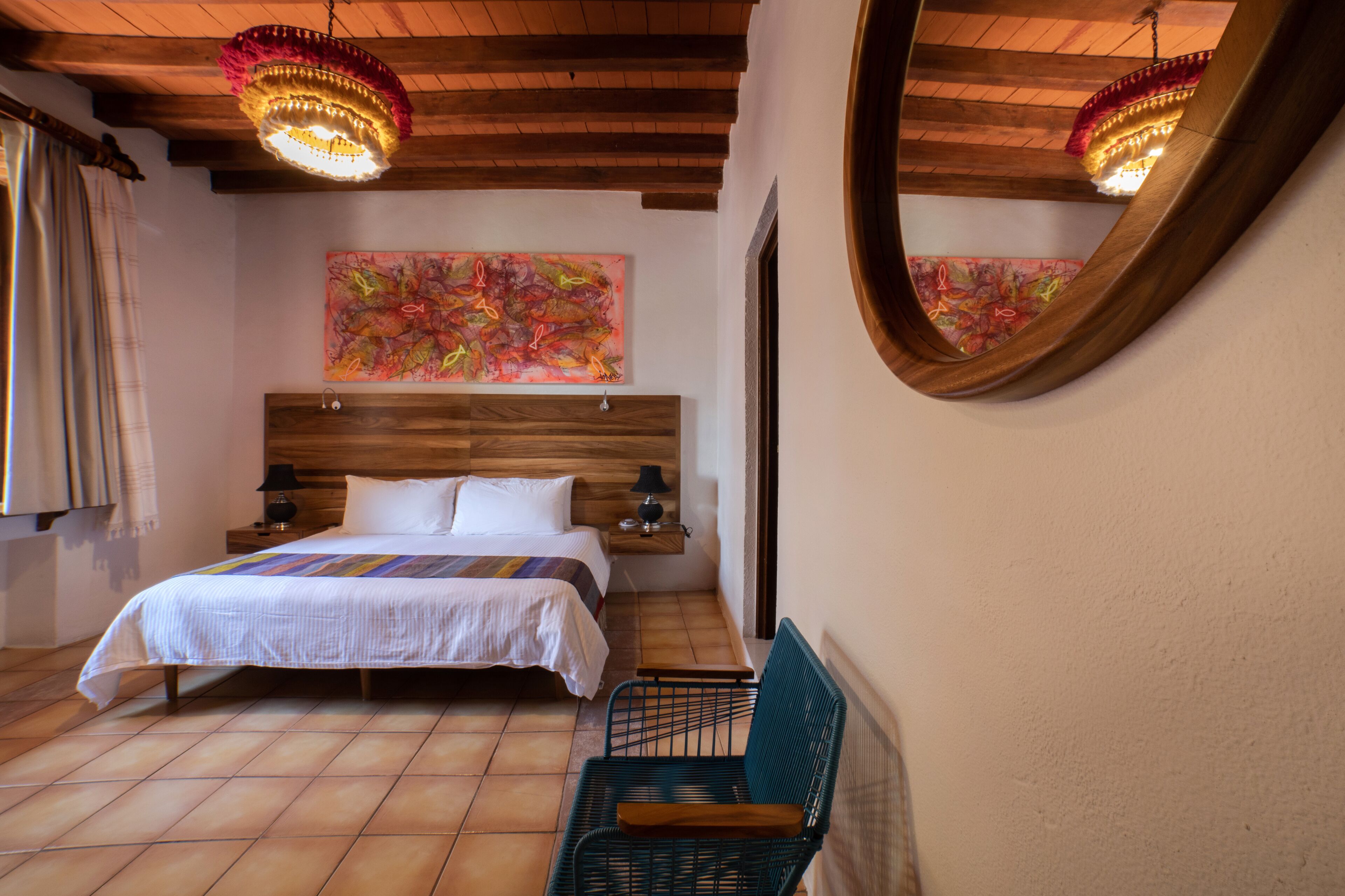 Un dormitorio con cabecera de madera, cama con sábanas blancas, una silla verde, mesita de noche de madera, un espejo y un cuadro abstracto de colores en la pared.