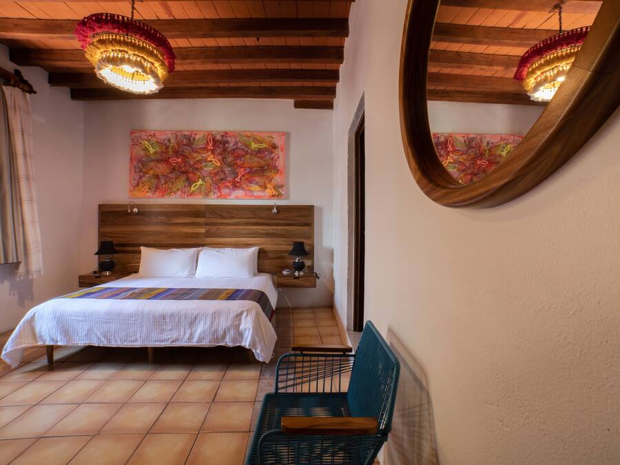 Un dormitorio con cabecera de madera, cama con sábanas blancas, una silla verde, mesita de noche de madera, un espejo y un cuadro abstracto de colores en la pared.