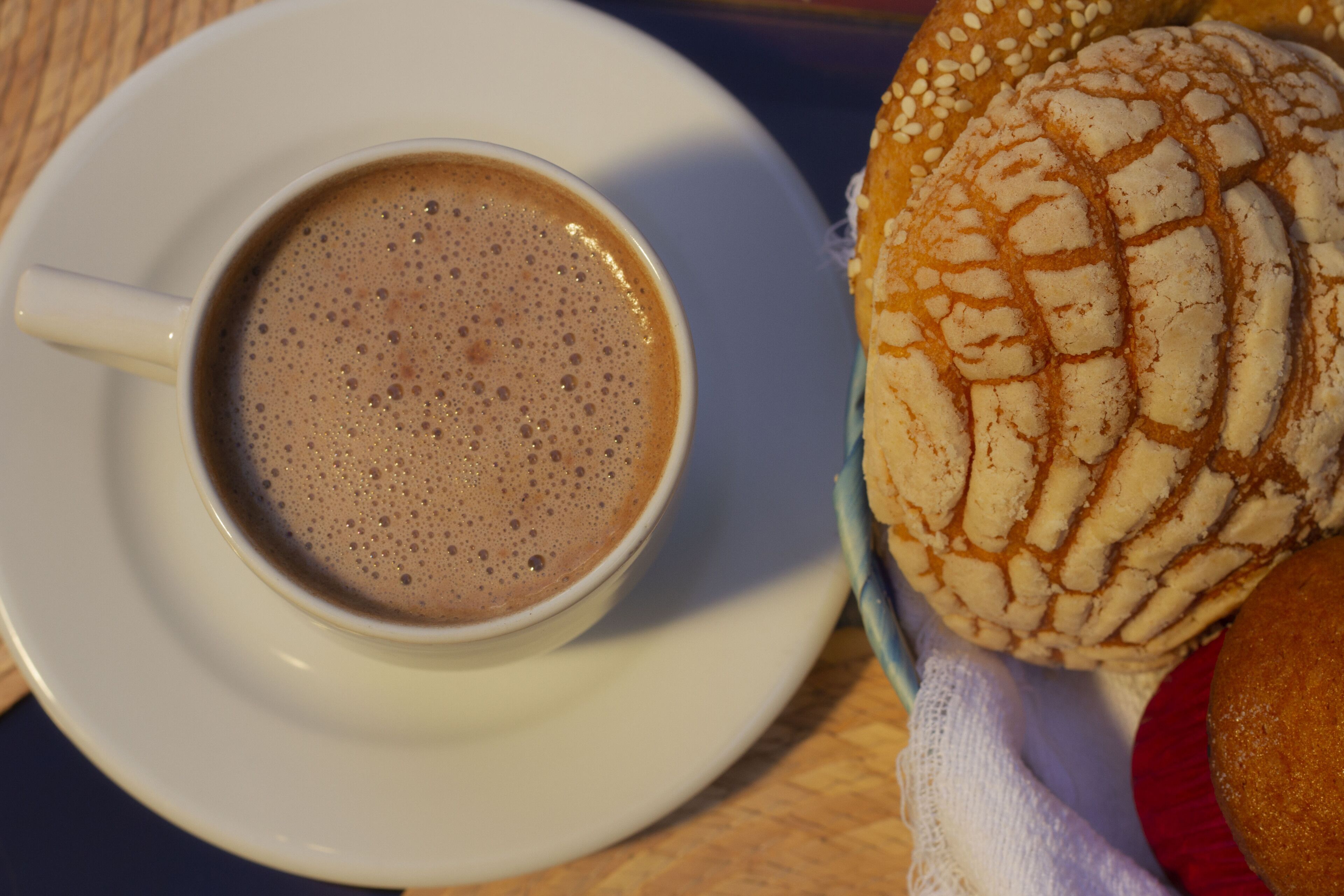 A cup of coffee on a saucer, a bread roll with sesame seeds, and a red object on a table.