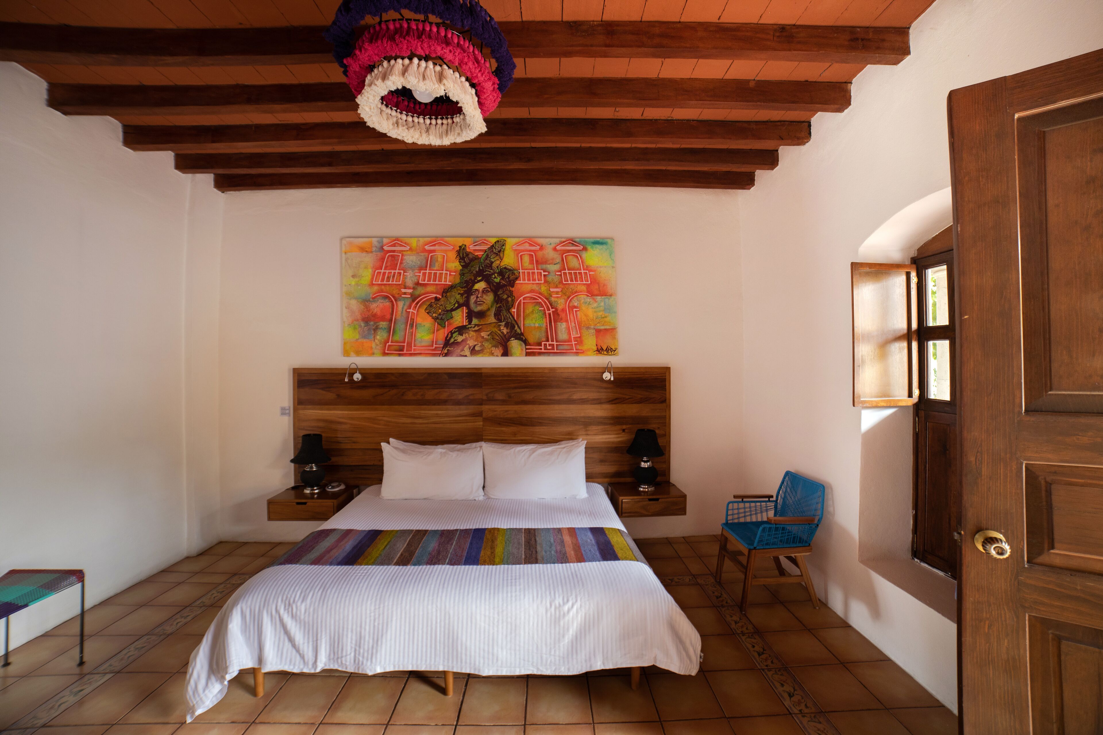 Ein Schlafzimmer mit einem Holz-Kopfteil, einem farbenfrohen Gemälde über dem Bett, einem blauen Sessel und einer Holztür.
