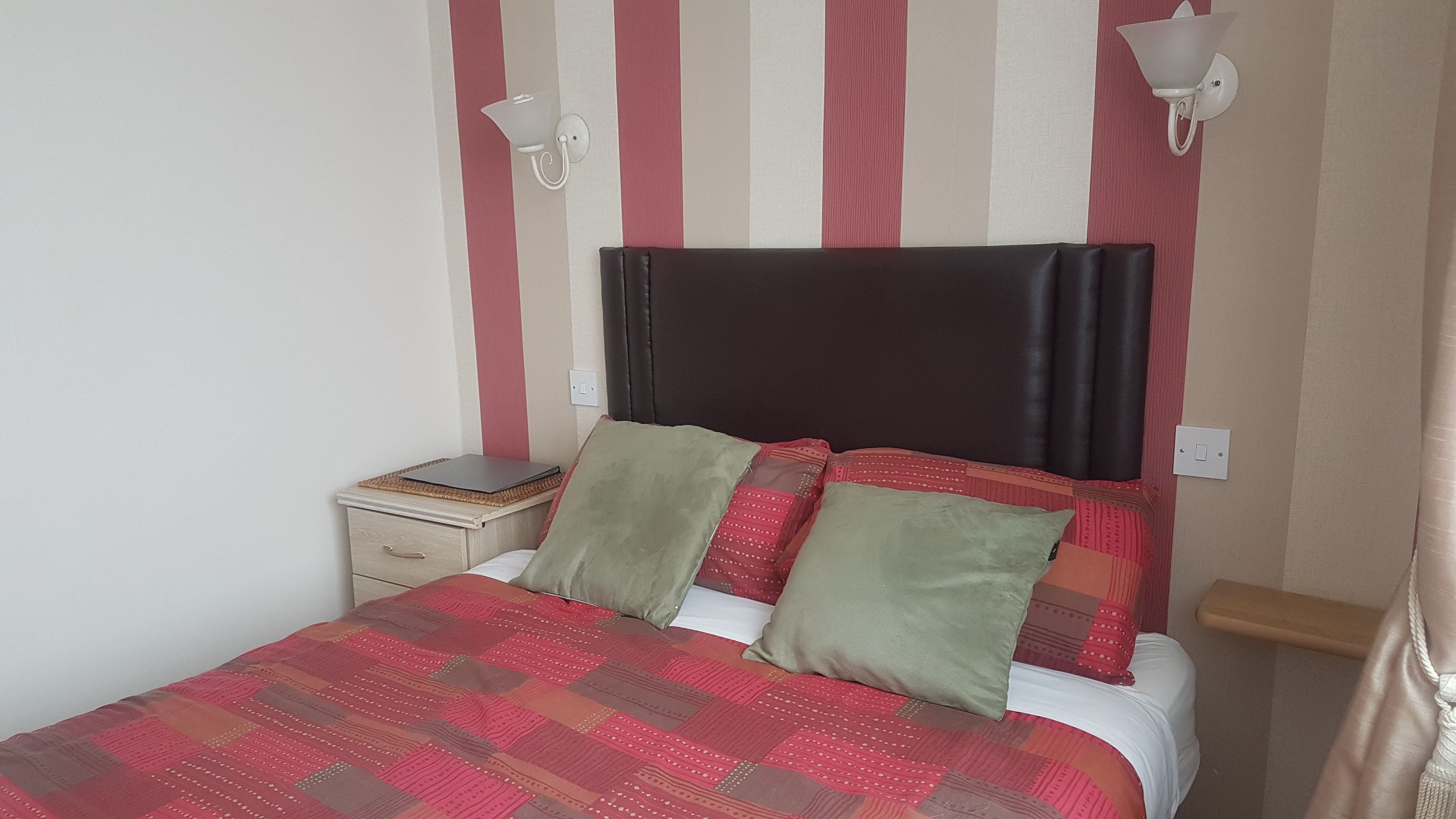 A bed with a red and brown quilt, two pillows, a nightstand, and a wall-mounted lamp.