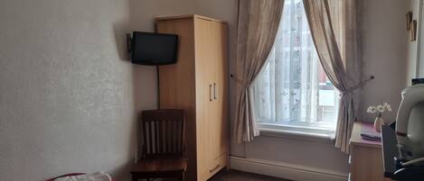 A small room with a wooden wardrobe, a single chair, a bed with a red and black checkered blanket, a television mounted on the wall, and a window with sheer curtains.