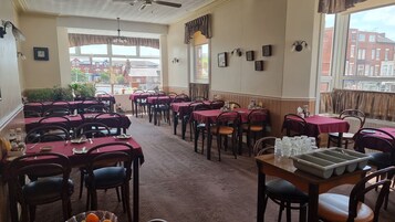 Een restaurant met meerdere tafels en stoelen, grote ramen met gordijnen en uitzicht op gebouwen.
