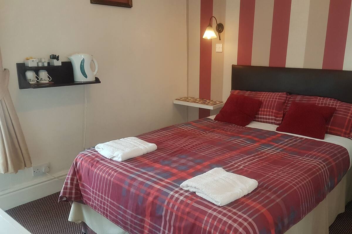 A neatly made bed with a plaid bedspread and two white towels, a bedside table with a lamp, and a shelf with kitchen items.