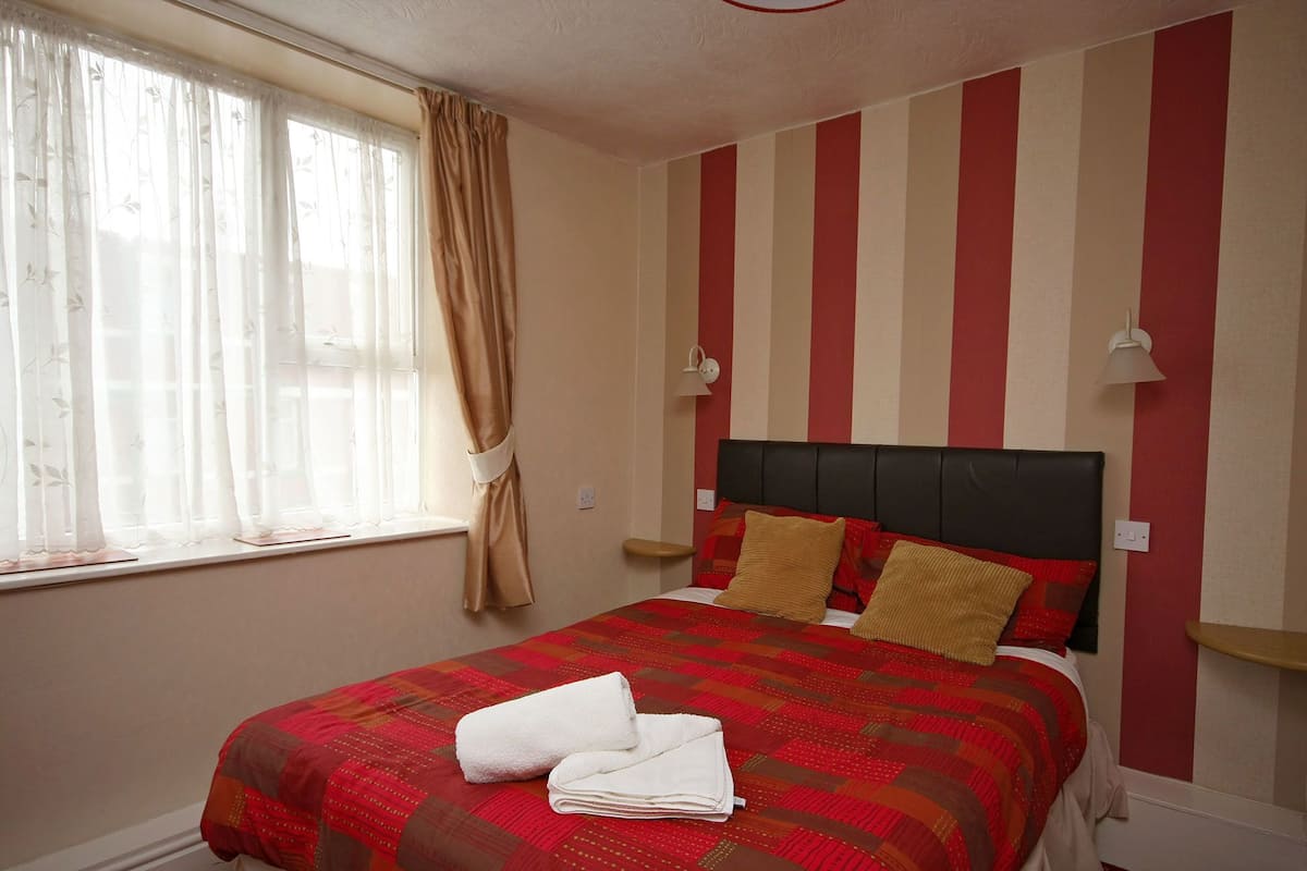 A neatly made bed with a red and brown quilt, two pillows, and a folded white towel on the bed.
