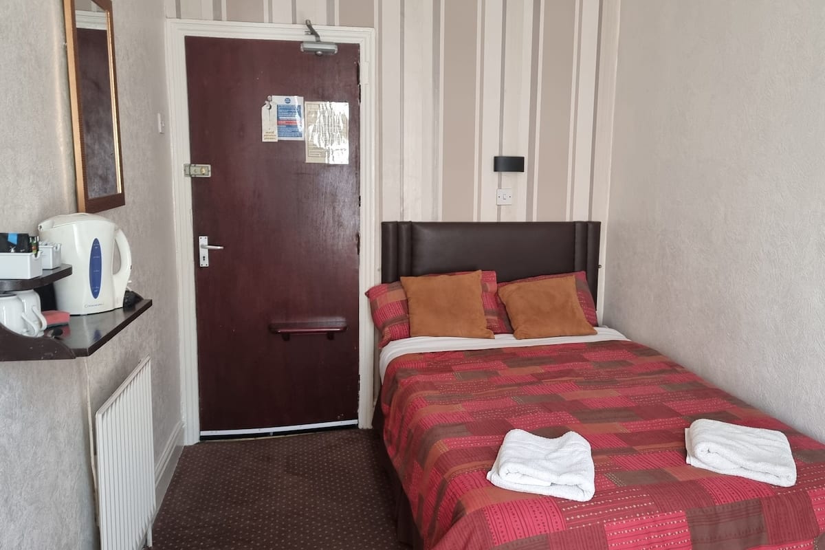 A small hotel room with a single bed, a nightstand with a kettle, a mirror, and a door with a notice.