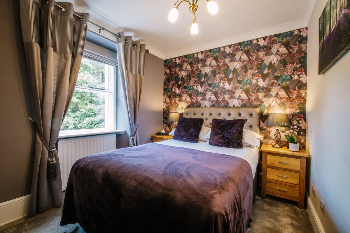 A bedroom with a large bed, a headboard with a lamp, a nightstand, and a floral-patterned wall.
