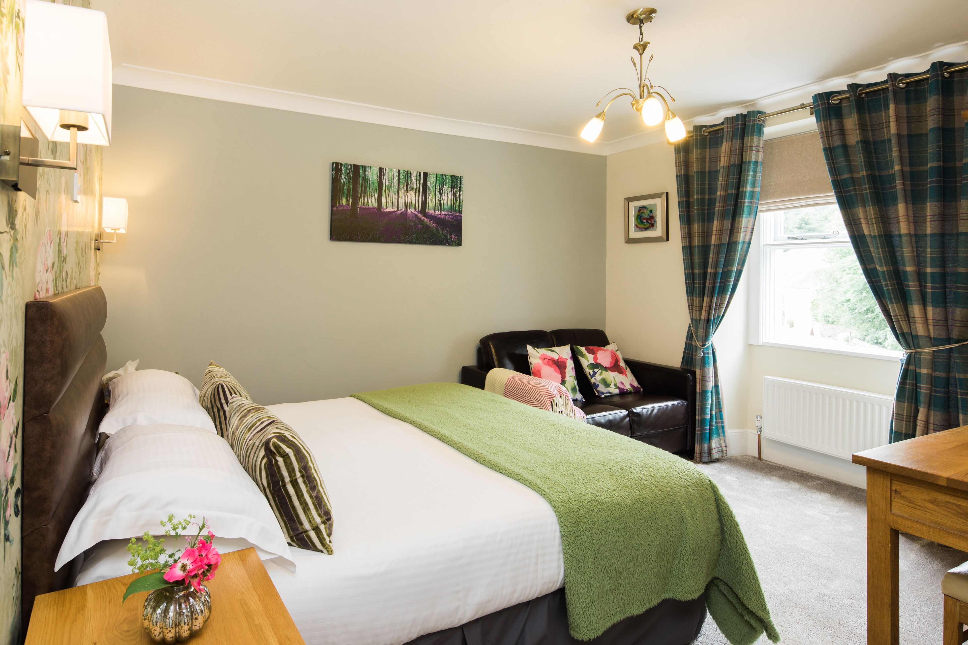 Une chambre à coucher avec un lit, une table de chevet en bois, un plaid vert, un vase avec des fleurs, un tableau représentant une forêt, un canapé en cuir noir, des rideaux à carreaux et une fenêtre donnant sur un paysage.