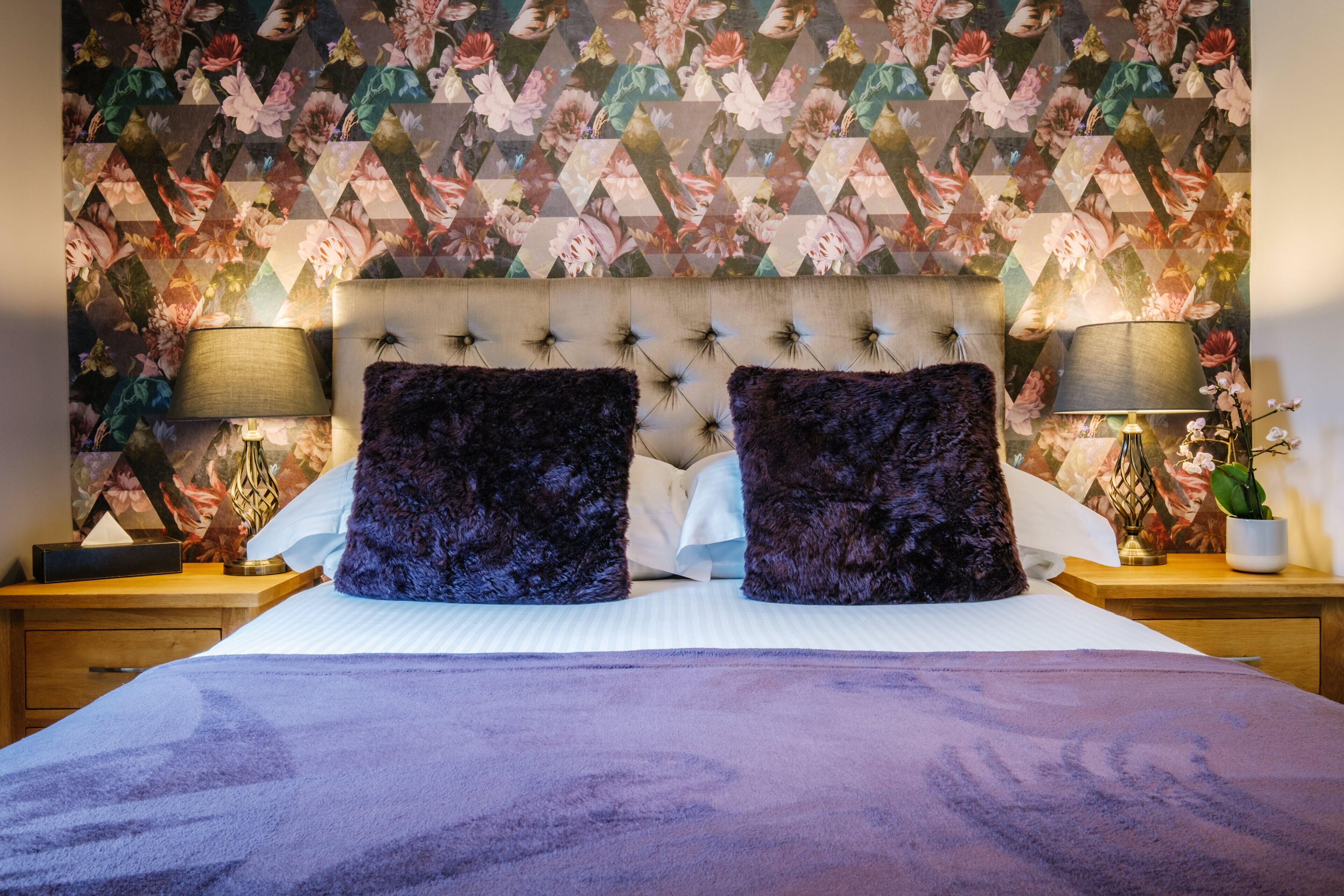 Un dormitorio con papel tapiz estampado con motivos florales, cabecera capitonada, dos almohadas moradas oscuras y lámparas de noche.