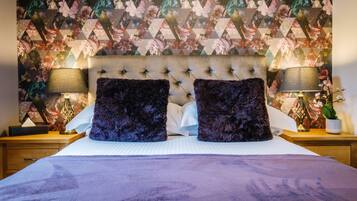 Un dormitorio con papel tapiz estampado con motivos florales, cabecera capitonada, dos almohadas moradas oscuras y lámparas de noche.