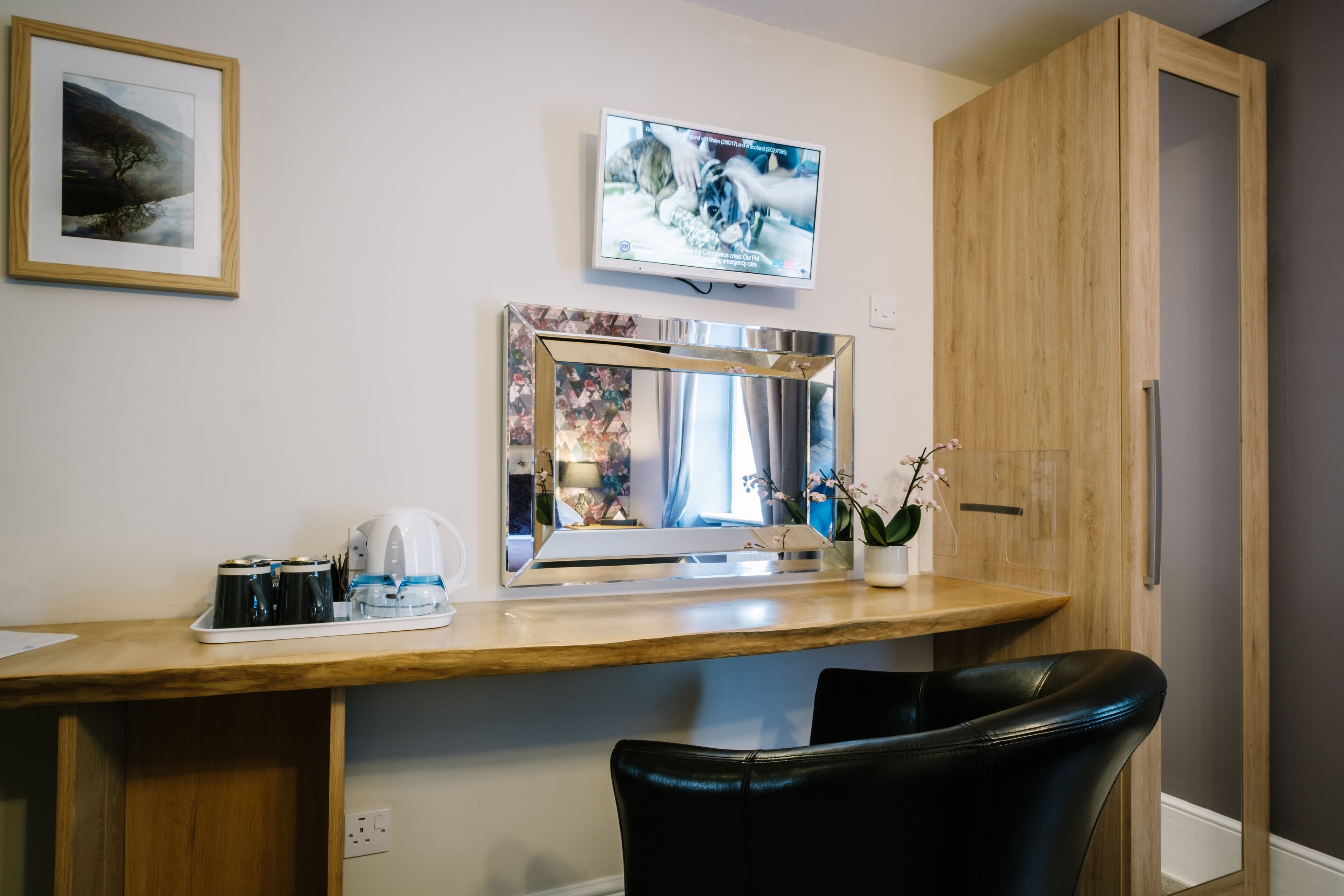 Une chambre d’hôtel avec un bureau en bois, un téléviseur à écran plat fixé au-dessus, un tableau encadré au mur et une orchidée en pot posée sur le bureau.