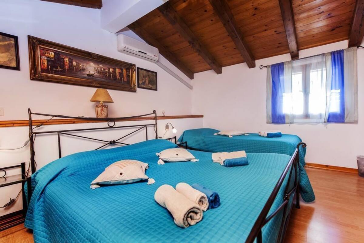 A hotel room with a wooden ceiling, a bed with blue bedding, a lamp, and a window with blue curtains.