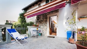 Ein Steinhaus mit einer gefliesten Terrasse, einem blauen Liegestuhl, einem kleinen Tisch und Topfpflanzen.