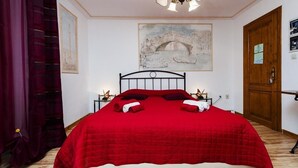Ein ordentlich bezogenes Bett mit roter Bettdecke und Kissen, Holzboden, ein Bild einer Brücke an der Wand und eine Holztür.