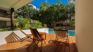 A wooden deck with two wooden chairs and a table, overlooking a swimming pool and lush greenery.