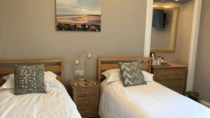 Quarto de hotel com duas camas de madeira, uma mesa de cabeceira com abajur, um quadro na parede e um espelho.