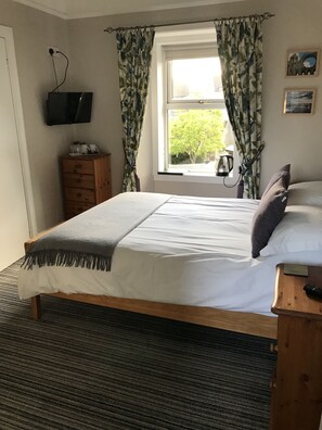 A bedroom with a bed, a wooden nightstand, a television mounted on the wall, and a window with curtains.