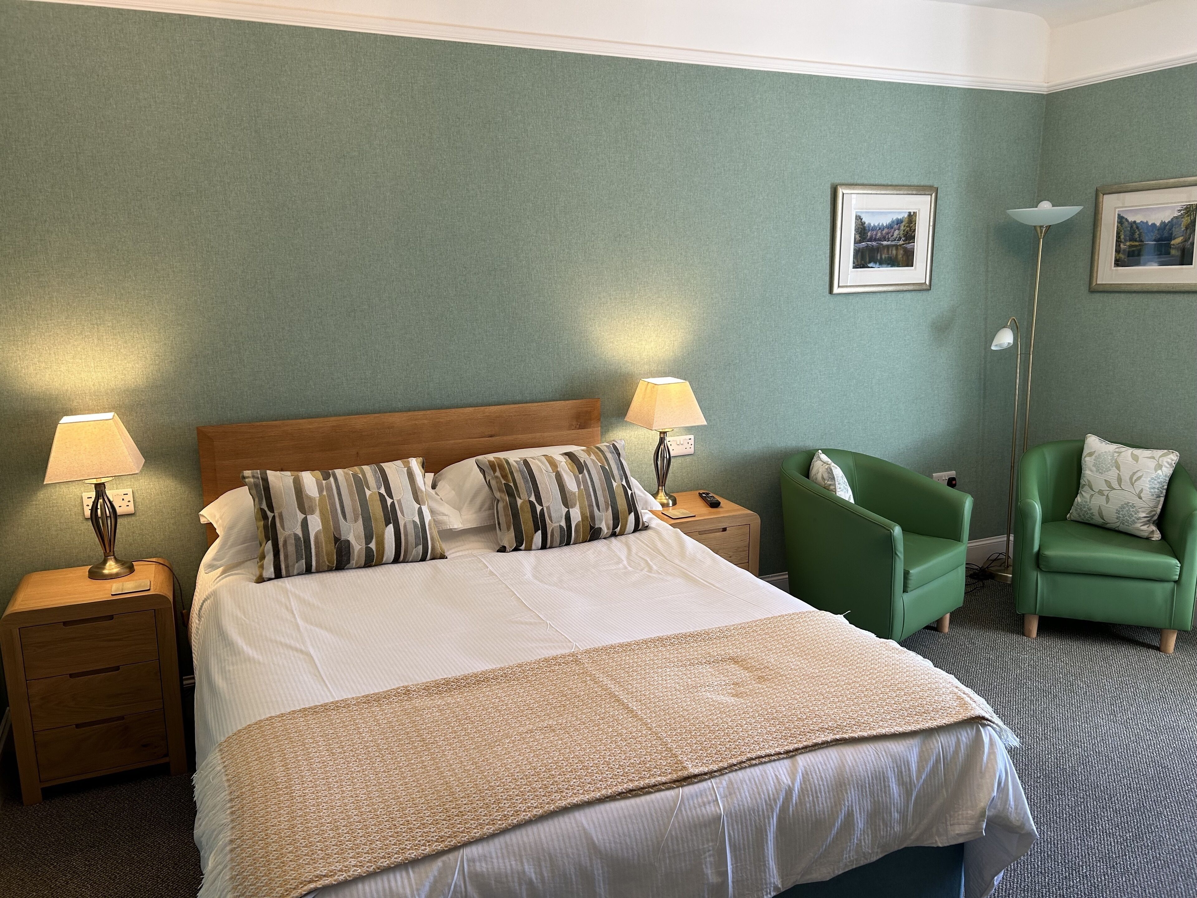 A hotel room with a large bed, two bedside tables with lamps, a green armchair, and two framed pictures on the wall.