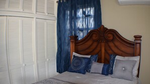 Une chambre à coucher avec une tête de lit en bois, un lit avec une literie  à motifs, une armoire aux portes blanches et des rideaux bleus.