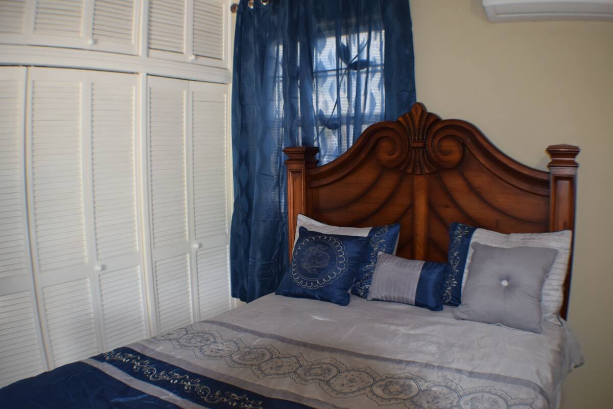 A bedroom with a wooden headboard, a bed with patterned bedding, a closet with white doors, and blue curtains.