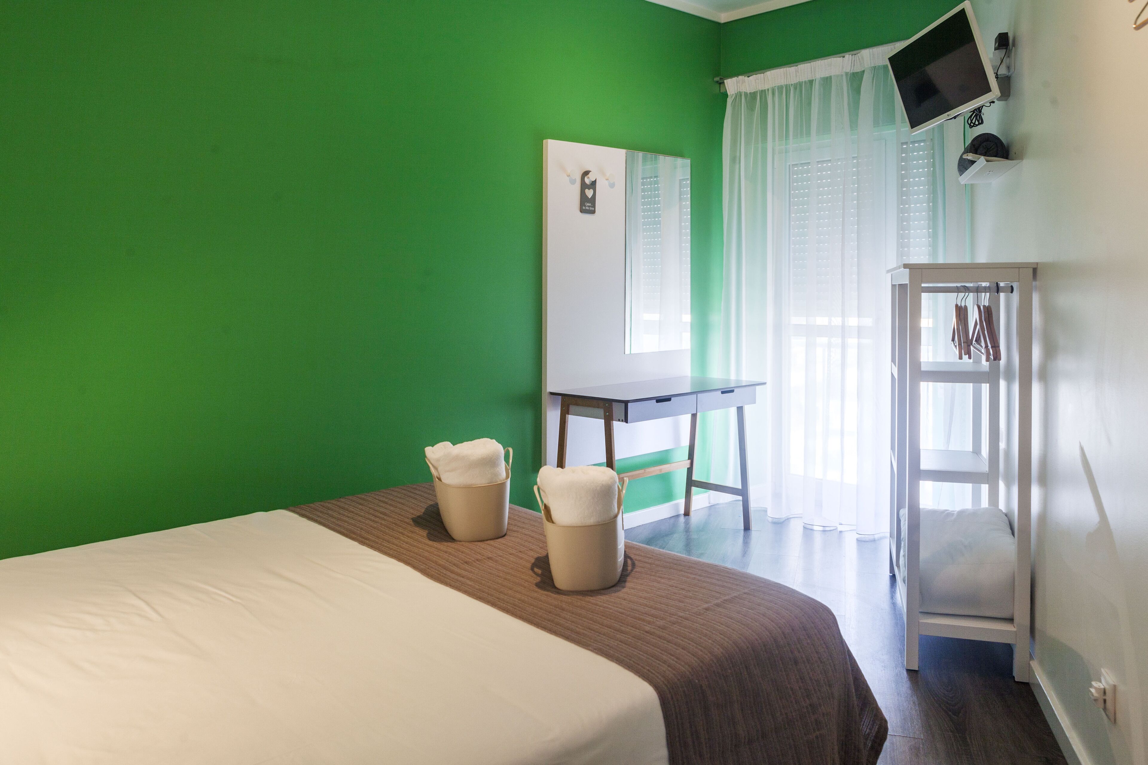 Une chambre d’hôtel avec un mur vert, un lit recouvert d’une courtepointe blanche et marron, deux tables de chevet beiges avec des serviettes, un bureau blanc avec un écran d’ordinateur et une armoire blanche.