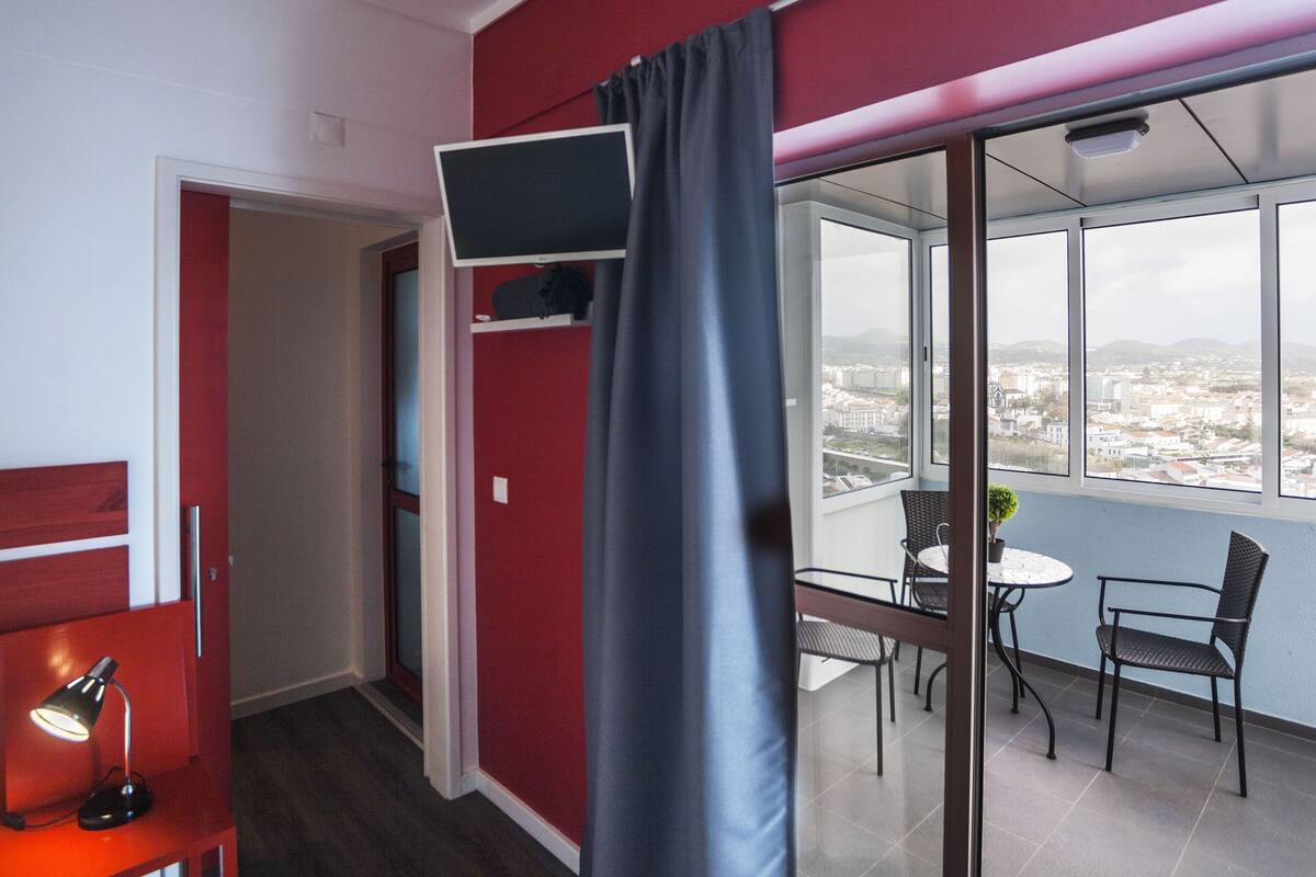 A modern room with a red accent wall, a flat-screen TV mounted on the wall, a balcony with a table and chairs, and a city view through large windows.